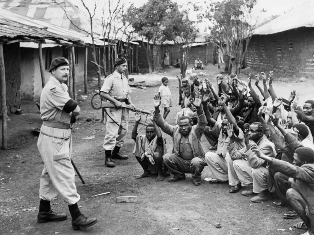 Mau Mau prisoners squatting with their hands up at gunpoint